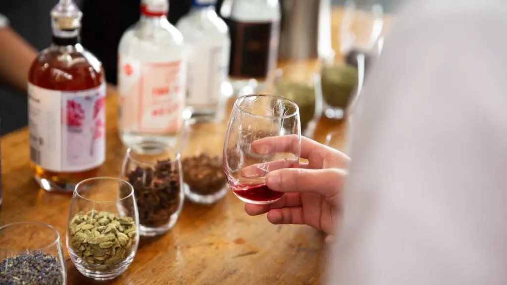 A hand holding a glass with red gin in focus, surrounded by various spices and bottles placed on a wooden table.