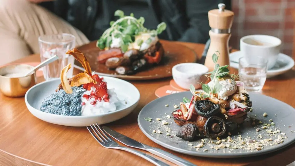A table with two plates of gourmet food, including dishes with various vegetables and garnishes, a glass of water, coffee, a pepper grinder, and utensils.