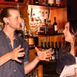 Two bartenders in denim shirts talk behind a bar stocked with bottles of liquor and wine; one holds a menu, both engaged in conversation during a cocktail making class in Melbourne.